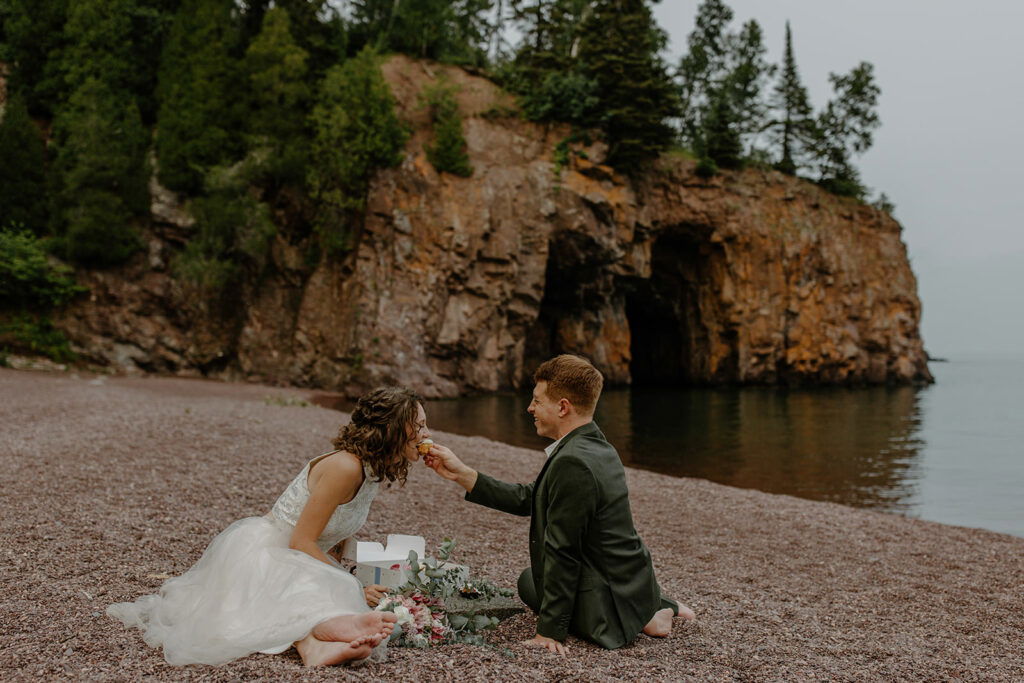 Couple in wedding attire sharing cake on a shore of pebbles and pine trees.