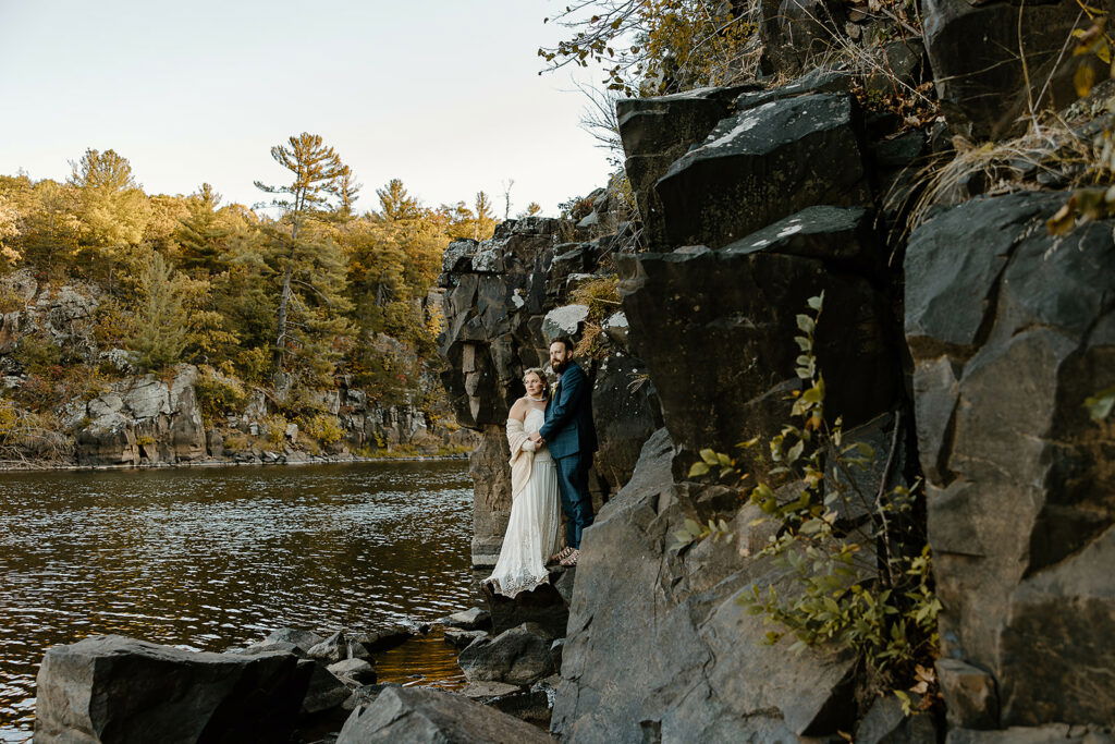 Couple in wedding attire standing on rocky cliff overlooking river.
