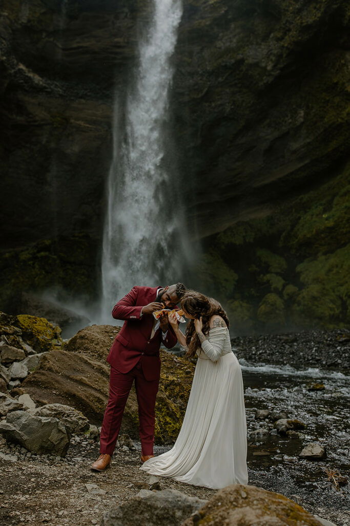 Couple in wedding attire eating hot dogs under a waterfall.